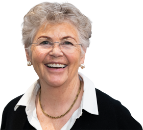 Lächelnde ältere Frau mit kurzen grauen Haaren, die eine Brille, Perlenohrringe, eine goldene Halskette, ein weißes Hemd mit Kragen und einen schwarzen Pullover trägt und vor einem schlichten hellen Hintergrund steht.