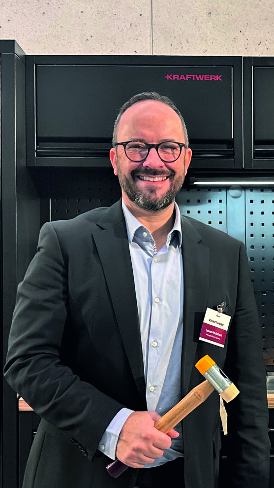 Un homme souriant portant des lunettes et un costume tient un marteau à tête jaune. Il se tient devant une unité de stockage d'outils noire étiquetée KRAFTWERK. Il porte un badge nominatif accroché à sa veste.