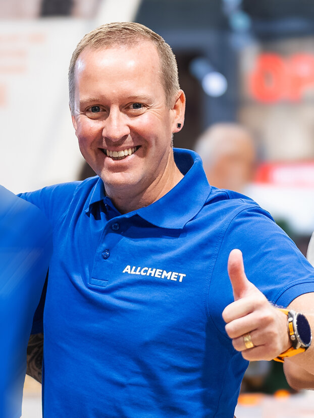 Eine lächelnde Person, die ein blaues ALLCHEMET-Poloshirt trägt, zeigt den Daumen nach oben. Sie hat kurze Haare, Ohrringe und eine Uhr und steht in einem Innenraum mit unscharfem Hintergrund.
