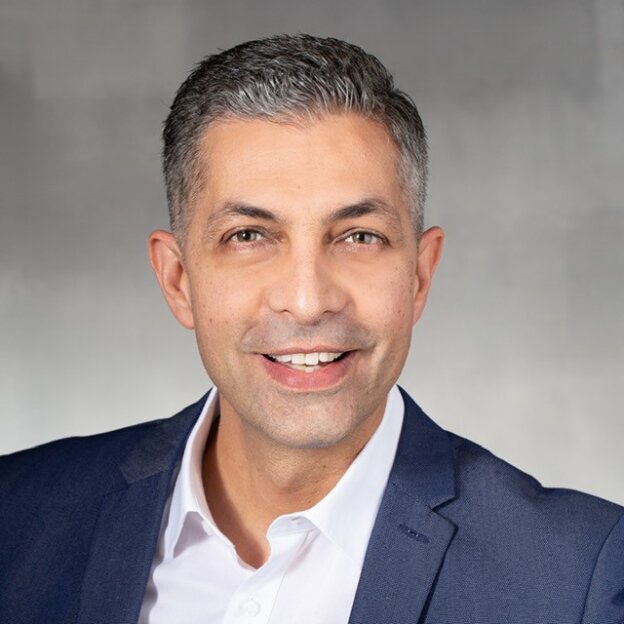 Ein lächelnder Mann mit kurzen grauen Haaren, der einen marineblauen Blazer und ein weißes Hemd trägt, posiert vor einem schlichten grauen Hintergrund.