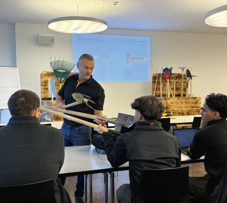 Ein Mann führt drei sitzenden Schülern in einem Klassenzimmer Gartengeräte vor, wobei hinter ihm Gartengeräte ausgestellt sind und eine Harke und eine Hacke auf einer Projektionsfläche gezeigt werden. Ein Schüler hält ein Werkzeug, während die anderen zusehen und sich Notizen machen.