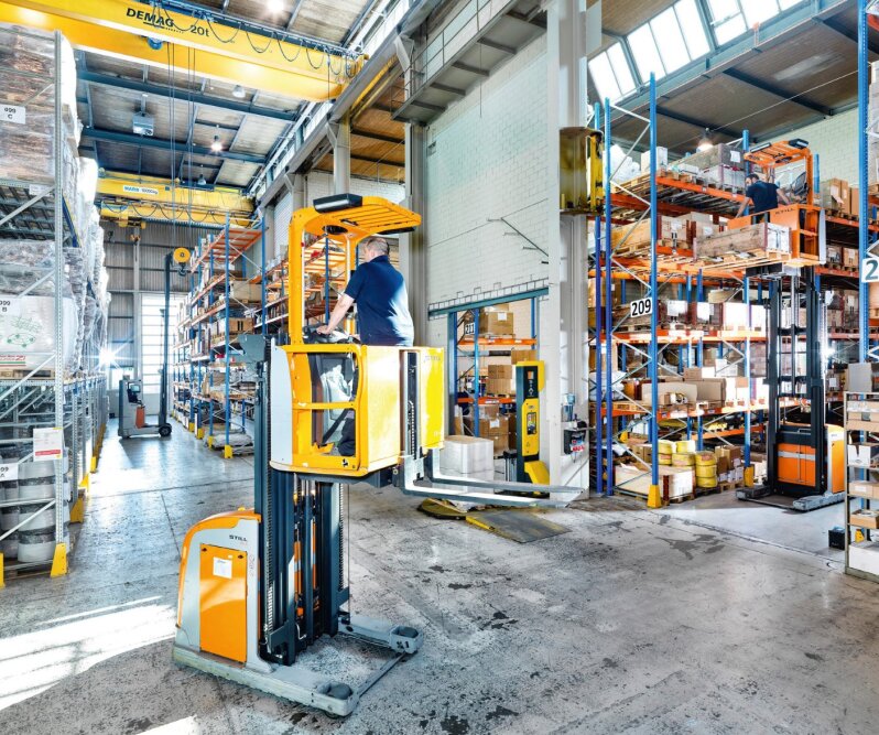 Ein Arbeiter bedient einen gelben Gabelstapler in einem hellen, gut organisierten Lager mit hohen Regalen, die mit verschiedenen Kisten und Gegenständen gefüllt sind. Das Sonnenlicht strömt durch hohe Fenster und erhellt die geräumige, geschäftige Umgebung.