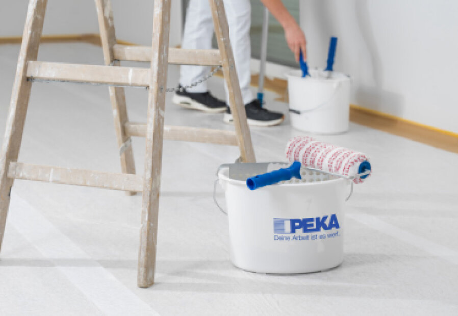 Eine Farbrolle ruht auf einem weißen Eimer mit der Aufschrift PEKA neben einer Holzleiter. Im Hintergrund bereitet sich eine Person in weißer Hose mit einer weiteren Rolle in einem separaten Eimer auf das Streichen vor.