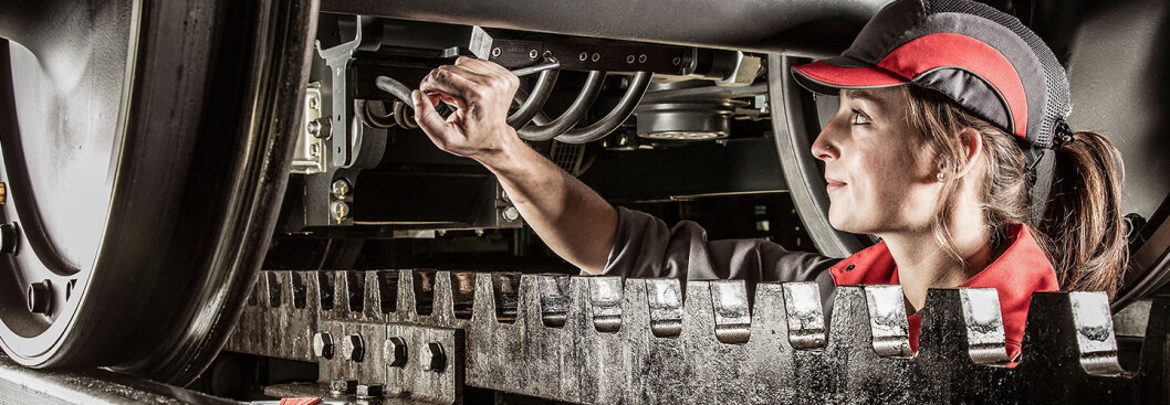 Eine Frau in rot-schwarzer Uniform und Mütze inspiziert oder repariert Maschinen unter einem großen Fahrzeug, wobei sie sich auf die Verkabelung und Komponenten konzentriert. Sie arbeitet mit Werkzeugen in einer industriellen Umgebung.