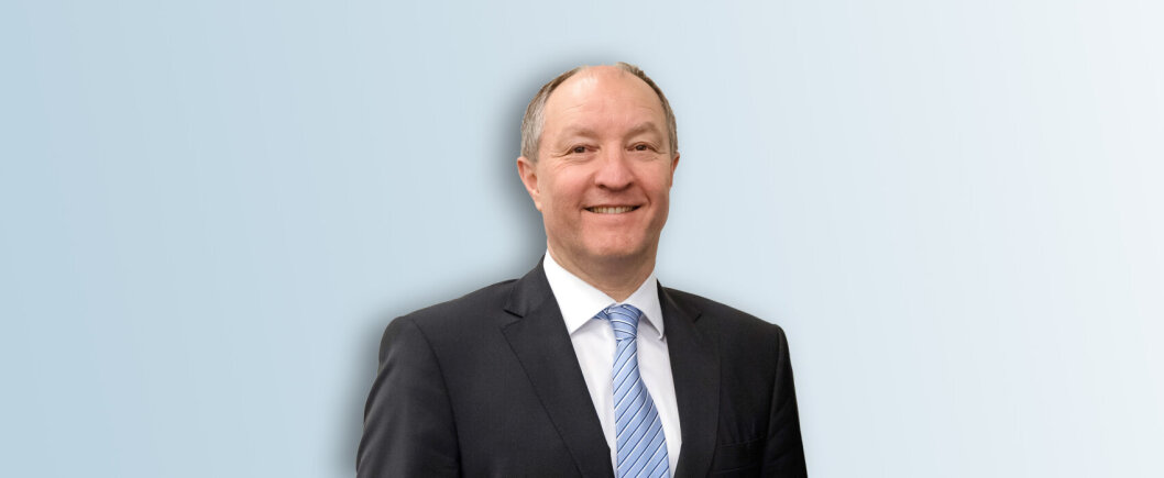 Un homme souriant, vêtu d'un costume sombre, d'une chemise blanche et d'une cravate rayée bleu clair, se tient sur un fond dégradé bleu clair.