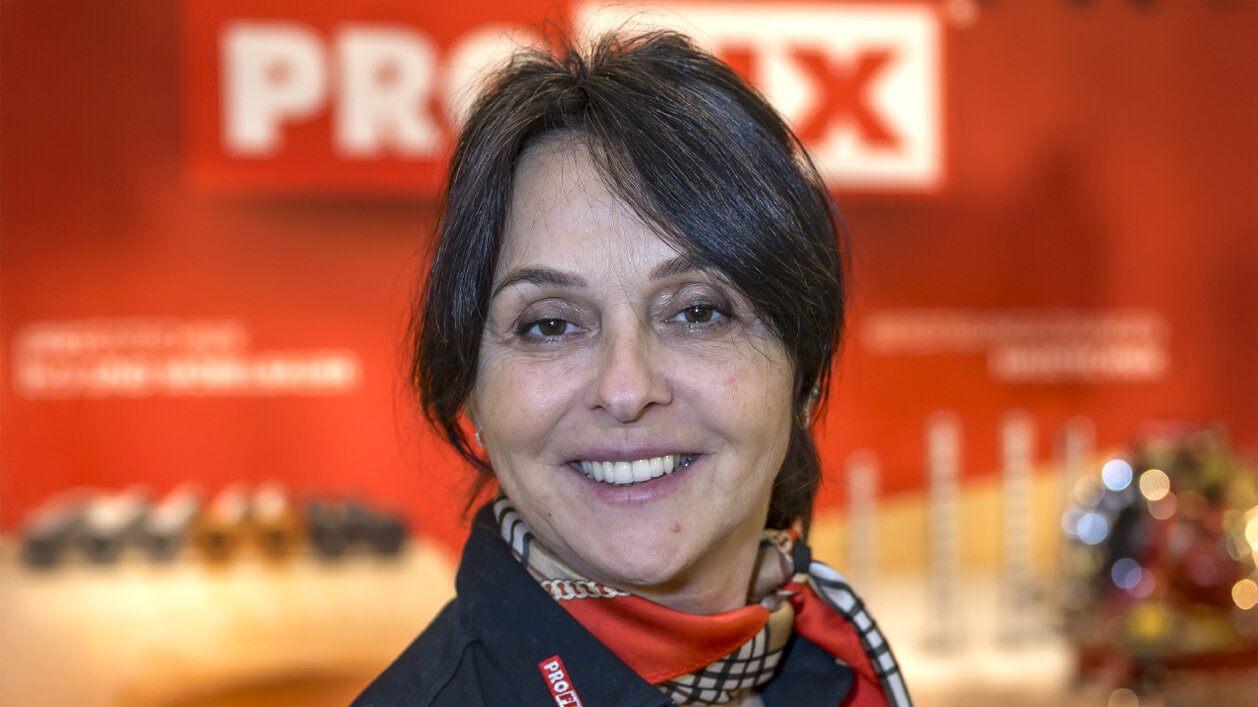 Eine Frau mit kurzen dunklen Haaren lächelt in die Kamera. Sie trägt eine schwarze Bluse und einen rot gemusterten Schal. Der Hintergrund ist rot mit unscharfem Text und Objekten, was auf ein berufliches Umfeld oder eine Veranstaltung hindeutet.