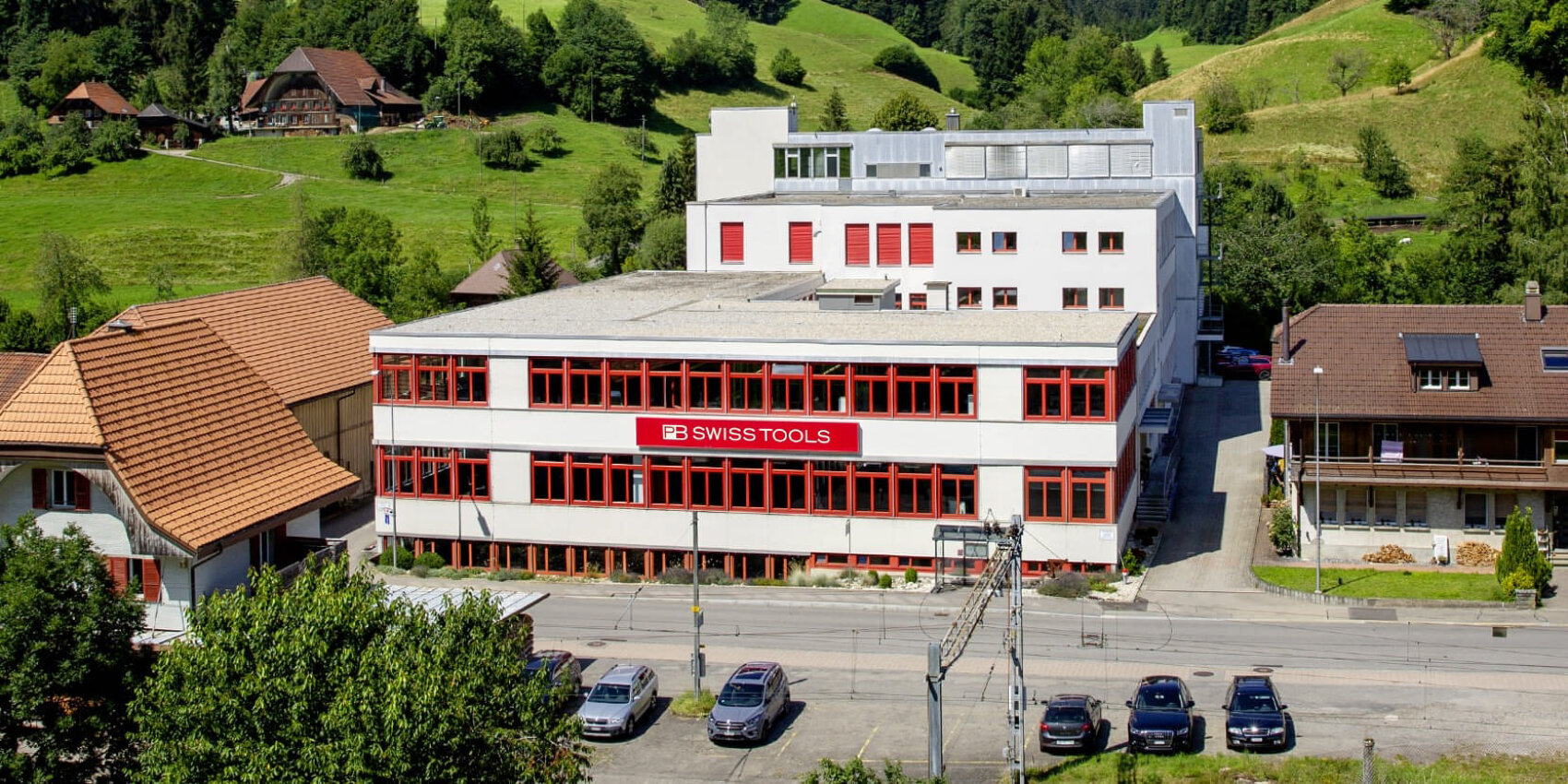 Ein großes weißes Gebäude mit rot umrahmten Fenstern und der Aufschrift PB SWISS TOOLS steht neben einer Straße mit parkenden Autos, umgeben von grünen Hügeln und verstreuten Häusern in einer ländlichen Gegend.