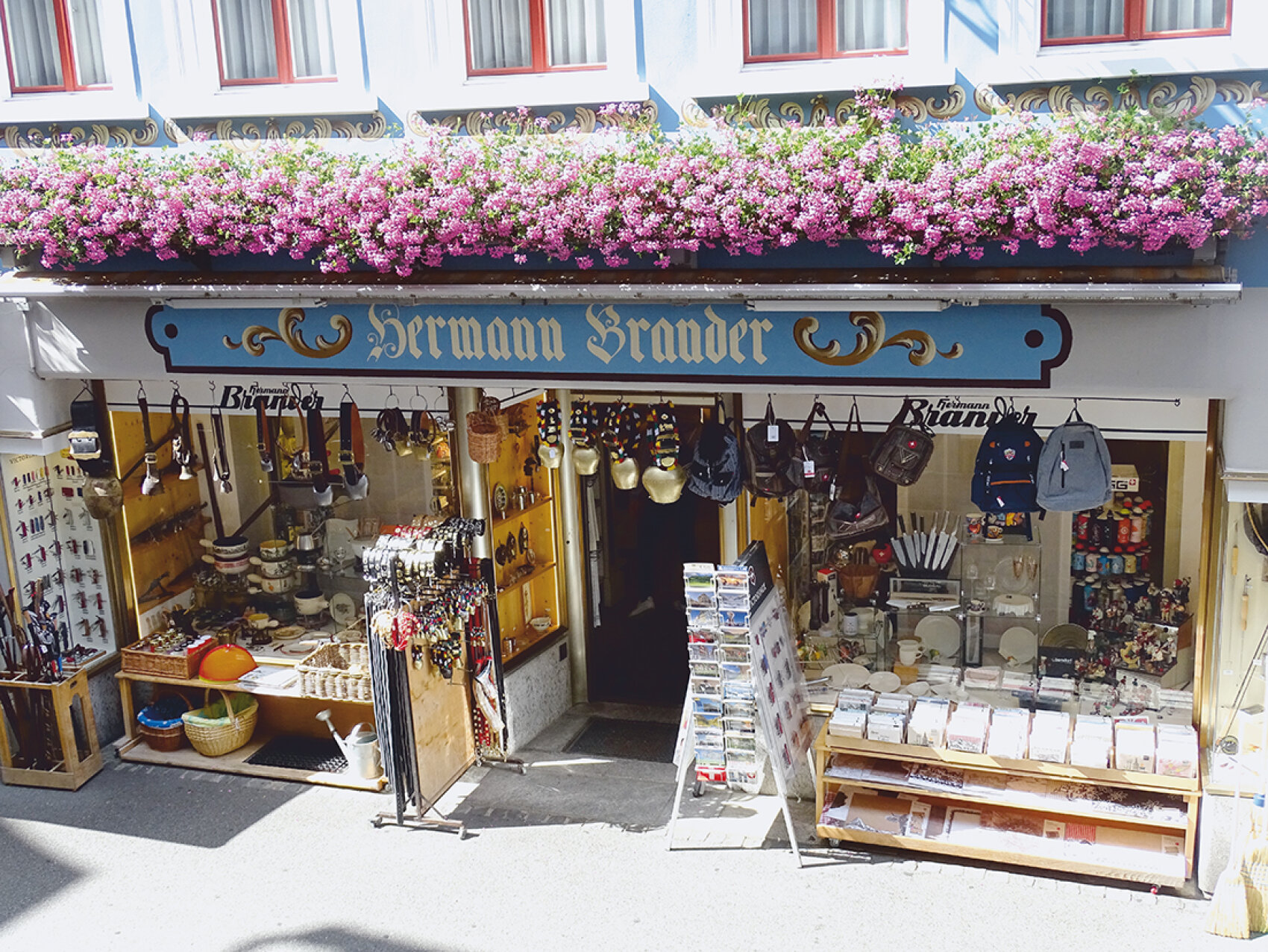 Une boutique pittoresque avec une enseigne bleue portant l'inscription Hermann Brauder, des fleurs colorées au-dessus de l'entrée et diverses marchandises comme des sacs, des ustensiles de cuisine et des cartes postales exposées à l'extérieur et à l'intérieur de la boutique.